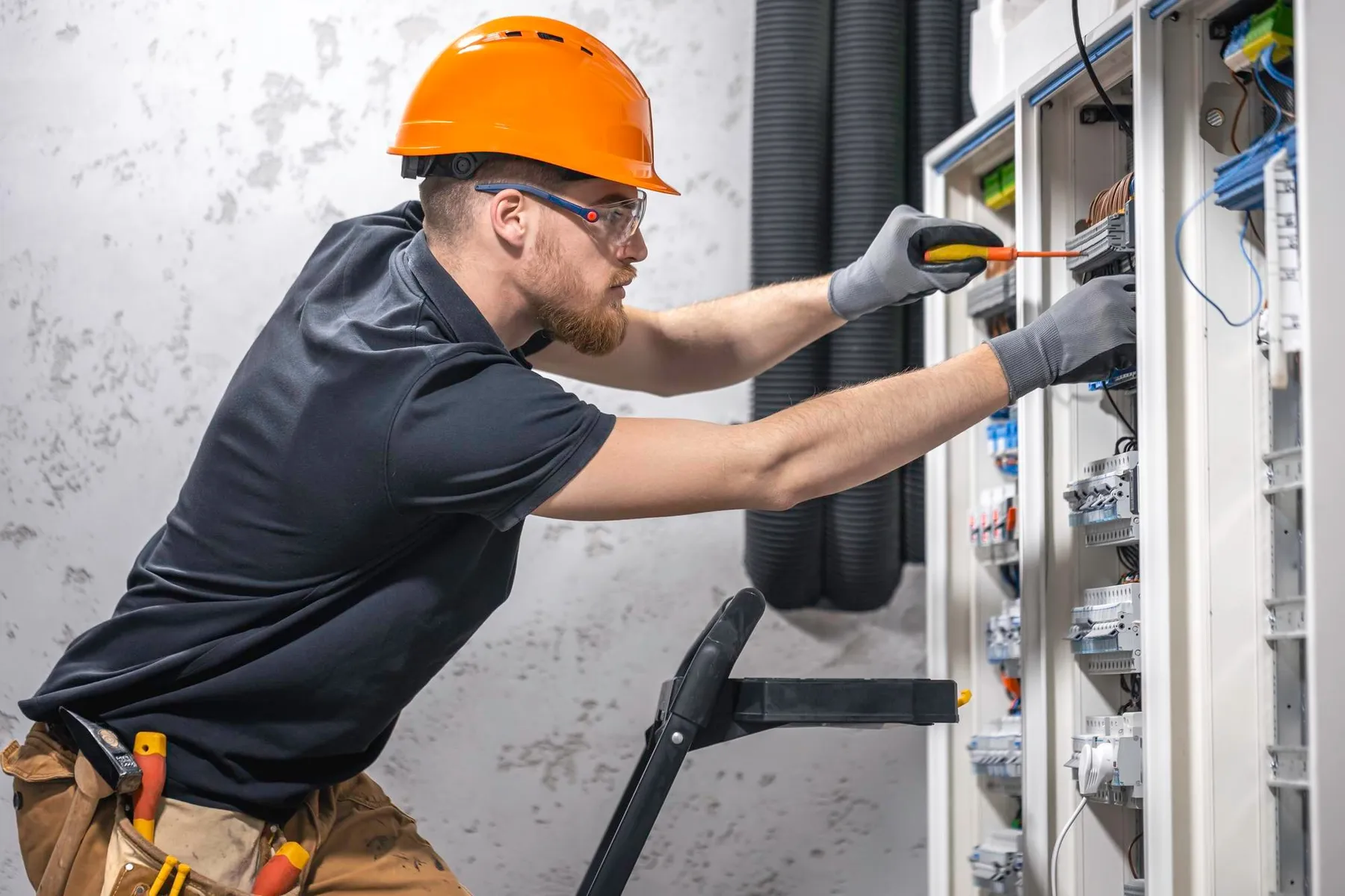 Villanyszerelő narancssárga sisakban, kesztyűben csavarhúzóval szerel egy nyitott elektromos kapcsolószekrényt.