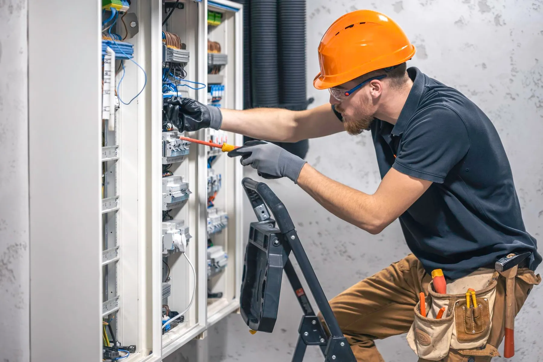 Villanyszerelő narancssárga sisakban, létrán állva vezetékeket és kapcsolókat szerel egy nyitott elektromos szekrényben.